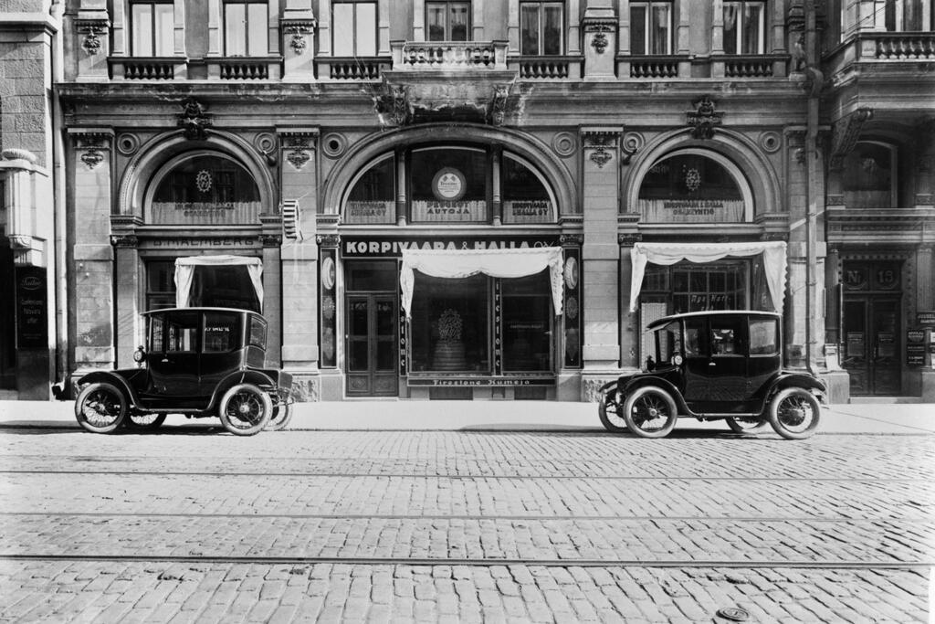 Autoja Aleksanterinkatu 15:ssa, taustalla Korpivaara & Halla Oy. Autot ovat Korpivaaran tuomia amerikkalaisia, sähkökäyttöisiä Rauch & Lang -merkkisiä. Hallan historiikin mukaan näitä tuotiin nämä kaksi kappaletta, sillä polttoaineesta oli pulaa sisällissodan kynnyksellä. Ajoneuvoja myytiin Helsingin Sanomissa Baker-merkkisinä vuonna 1920. Kuuluisa sarjakuvahahmo Mummo Ankka ajaa samantapaisella sähköautolla, mutta se on merkkiä Detroit Electric. Kuvaaja: Helsingin kaupunginmuseo / Eric Sundström