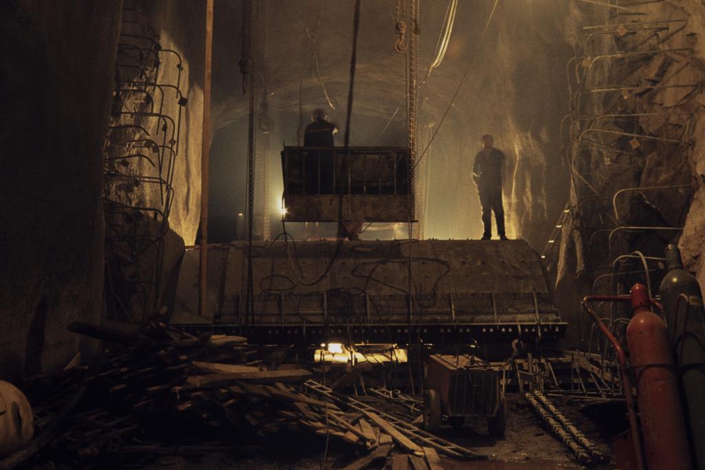 Kaksi työmiestä seisoo hämärässä, vielä rakenteilla olevassa metrotunnelissa. Toinen heistä seisoo maansiirtokoneen kauhassa.