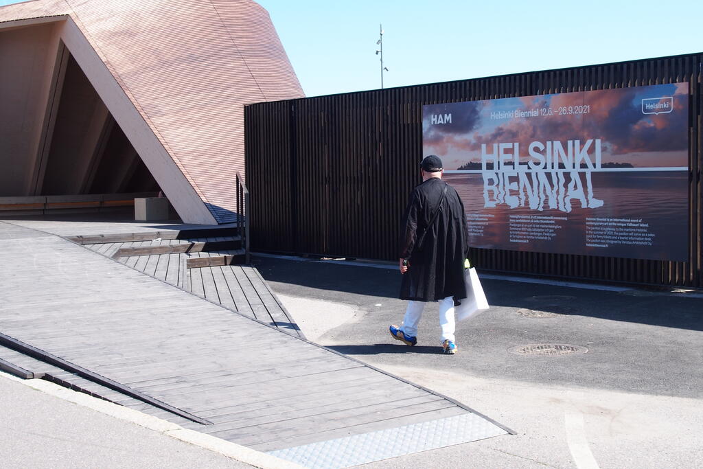 Helsinki Biennaalin mainos Etelärannassa. Etualalla takaapäin kuvattu, kävelevä ihminen.