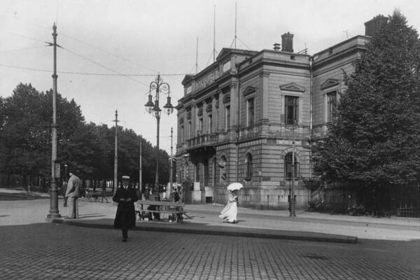 Vanhan ylioppilastalon edessä on koristeellinen kaasulyhty. Kadulla kävelee nainen, jolla on pitkä valkoinen mekko ja valkoinen päivänvarjo sekä mies, jolla on ylioppilaslakki.