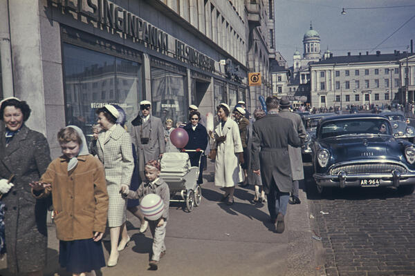 Människor med studentmössor promenerar på gatan. Södra kajen 18. I bildens högra kan en personbil av märket Buick och årsmodell 1954.