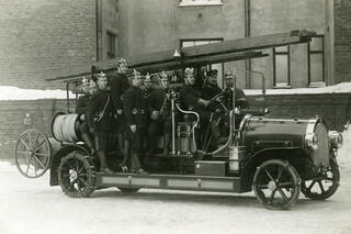Fyra brandmän ombord på en brandbil av öppen modell. De bär stövlar, uniformer och metallhjälmar.