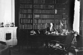 En man sitter bakom sitt skrivbord och har fotograferats framifrån. Bakom honom finns en bokhylla som täcker hela väggen och till vänster en vit kakelugn.