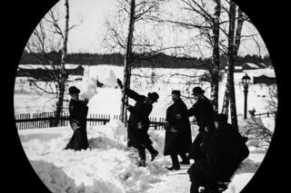 Hattuihin ja pitkiin takkeihin pukeutuvat miehet leikkivät lumisotaa pihatiellä. Taustalla näkyy koivuja, piha-aita ja kaasulyhty.