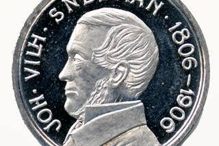 Keskellä hopeanväristä mitalia on miehen profiilikuva. Reunoja kiertää teksti JOH. VILH. SNELLMAN 1806–1906.