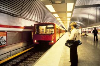 Metrotågen anländer till stationen. I förgrunden väntar en ung man i ylletröja med två väskor.
