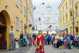 Etualalla jonglööri heittelee keiloja. Väkijoukko katselee taustalla, taka-alalla Tuomiokirkko.