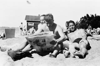 Uima-asuisia nuoria miehiä hiekkarannalla lukemassa sanomalehteä