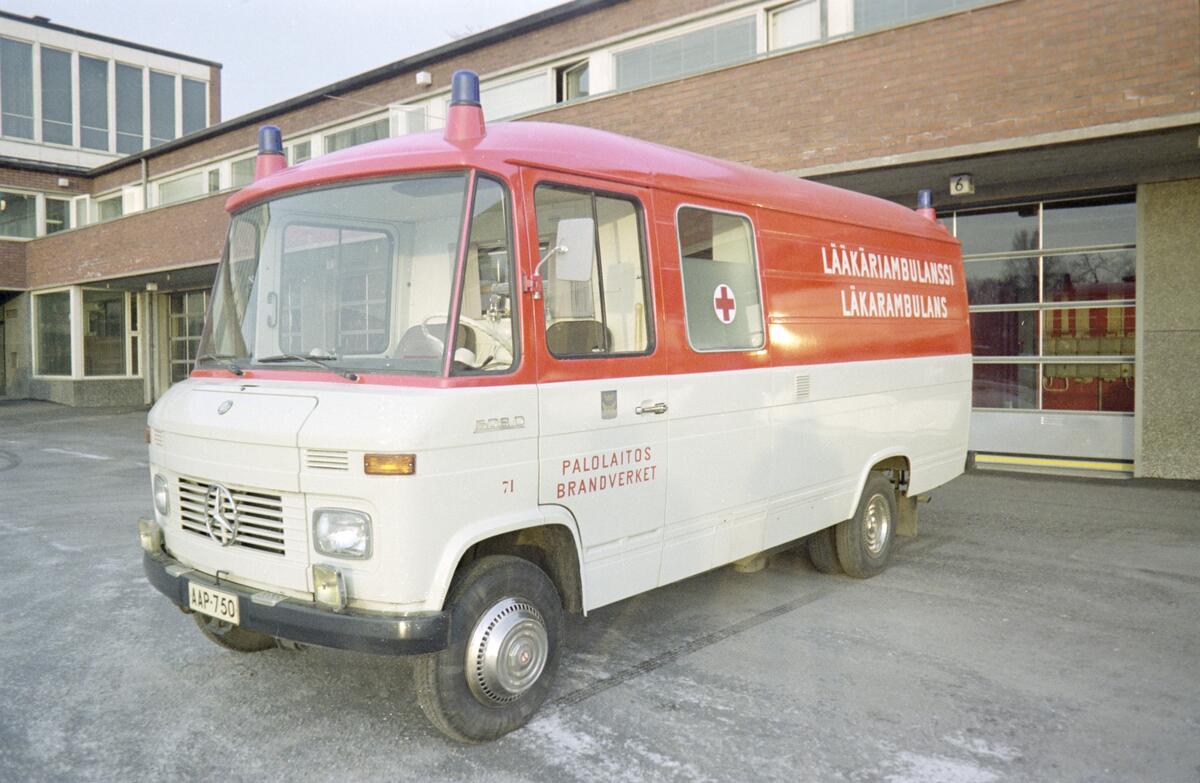 Ambulanssin alaosa on valkoinen ja yläosa punainen. Keulassa on Mercedes-merkki ja kyljissä teksti Lääkäriambulanssi Läkarambulans Palolaitos Brandverket.