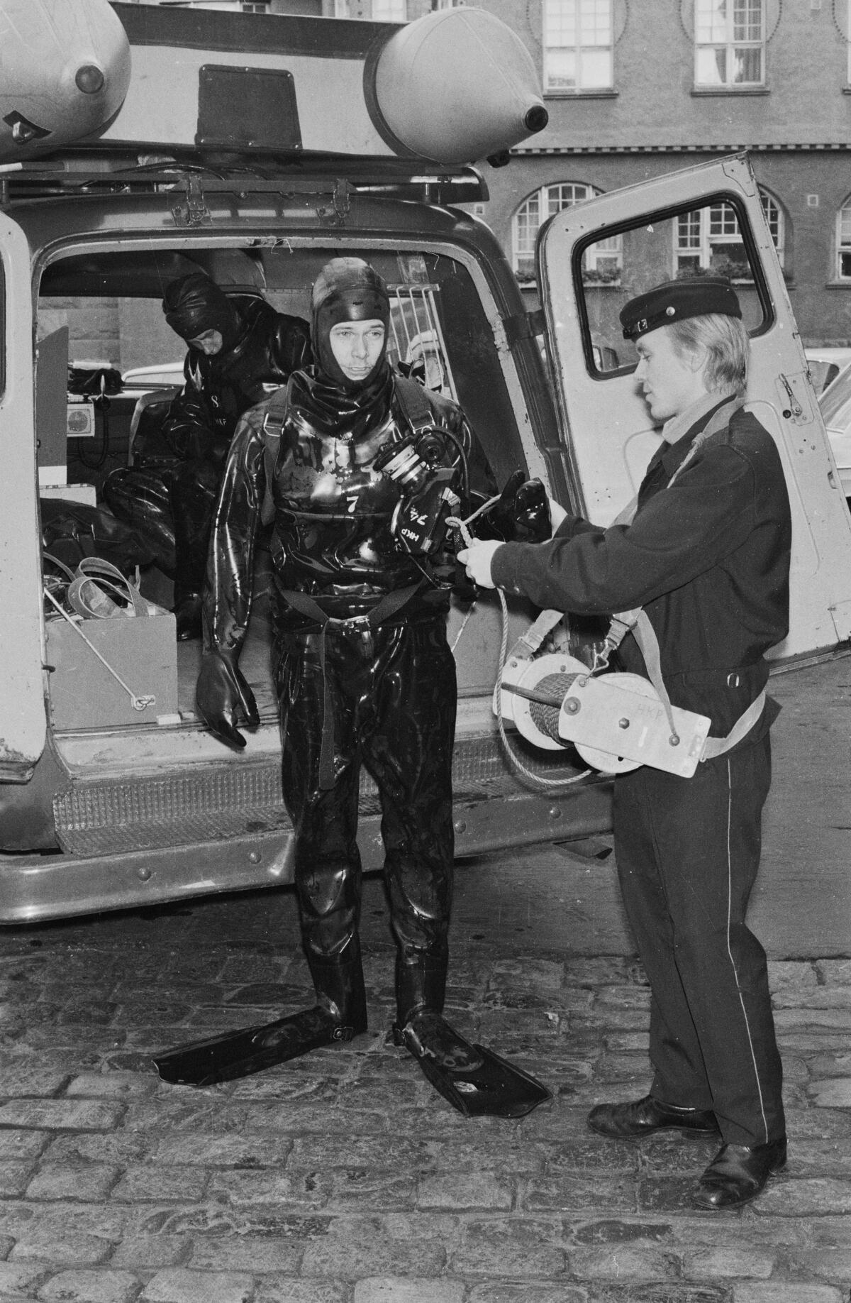 Pakettiauton avonaisten takaovien edessä seisoo sukelluspukuun ja räpylöihin pukeutunut mies. Hänen oikealla puolellaan seisoo sivuttain univormupukuinen mies. Auton sisällä näkyy osittain toinen sukelluspukuinen mies kyykyssä.