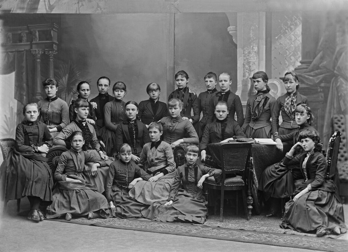 Pitkiin hameisiin ja korkeakauluksisiin puseroihin pukeutuneita nuoria naisia ryhmäkuvassa. Osa istuu lattialla, osa tuoleilla, osa seisoo.