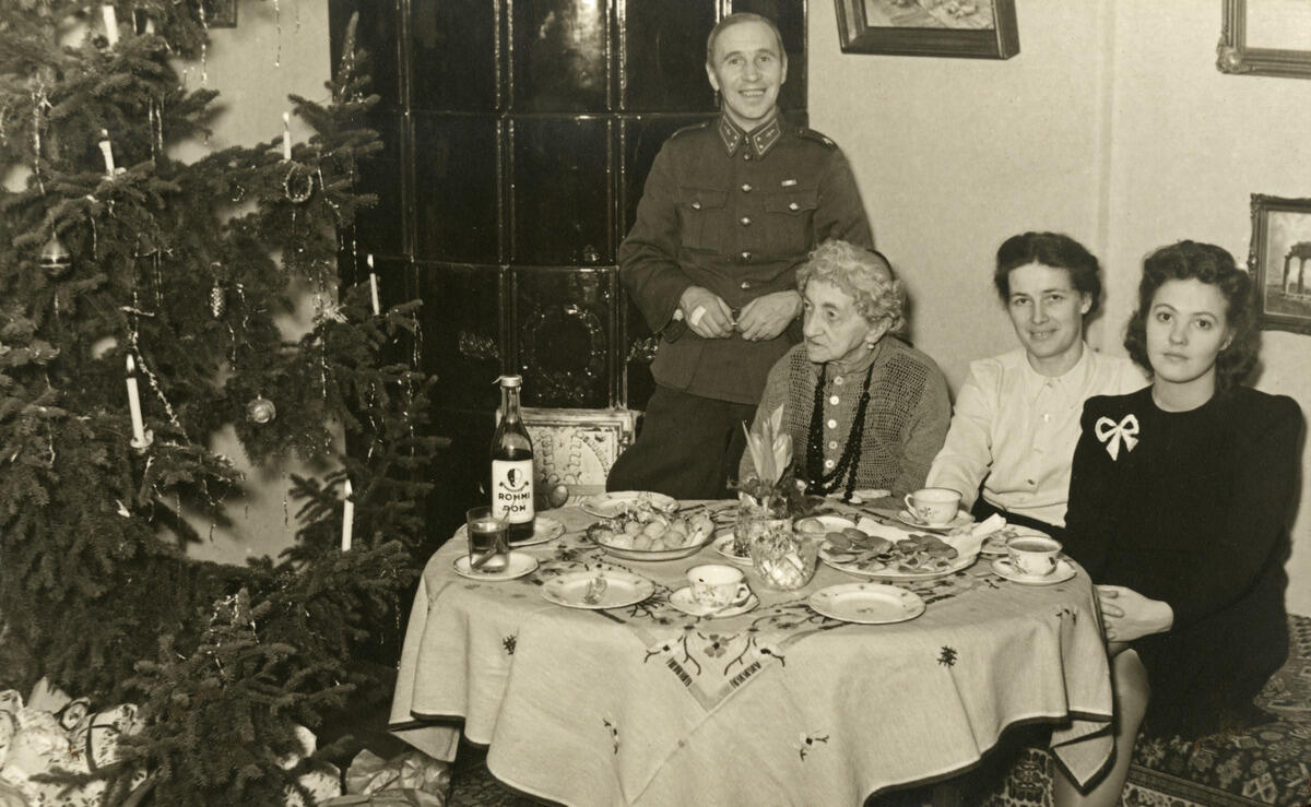 Joulunviettoa sota-aikana Haagassa (Vespertie 2). Vasemmalla on joulukuusi, jonka alla on lahjoja. Oikealla on kahvi- tai teepöytä, johon on katettu myös keksejä ja pullo rommia. Pöydän takana seisoo perheen isä sotilaspuvussa. Pöydän ääressä istuvat isän äiti, perheen äiti ja nuori nainen.
