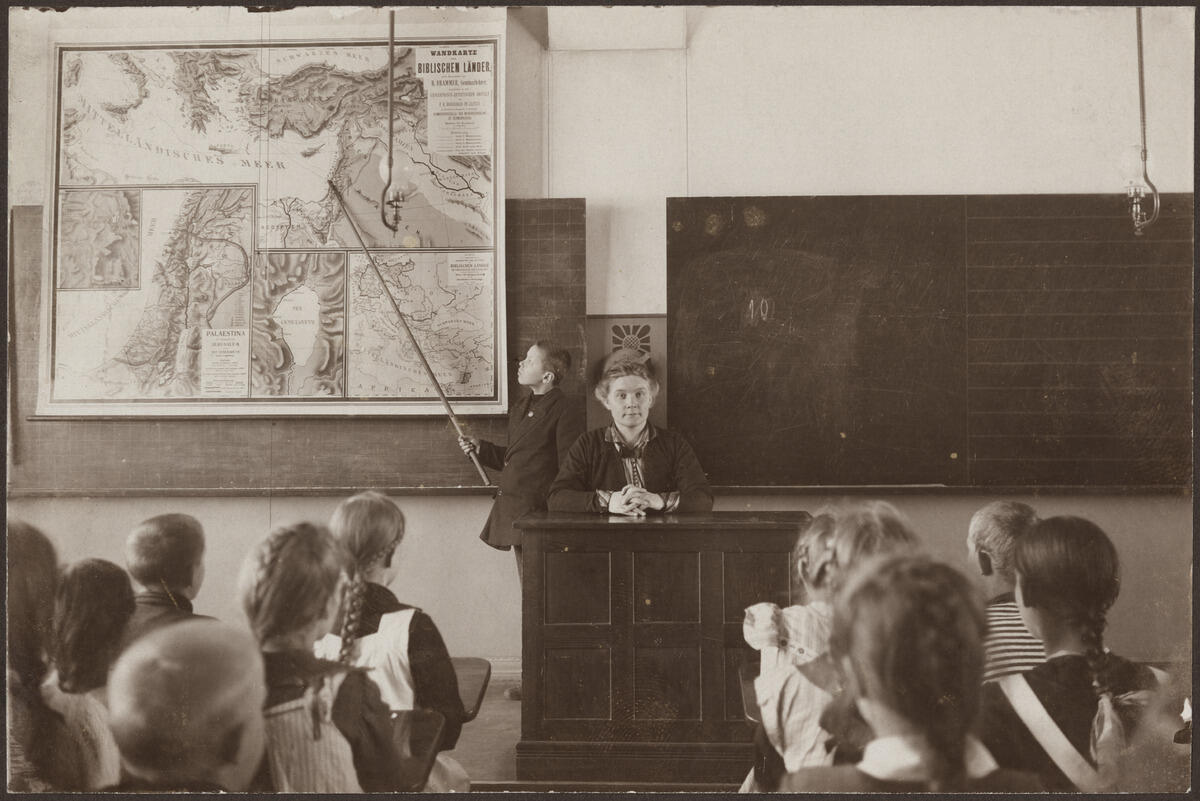 Oppilas näyttää Raamatun maita esittävältä kartalta Välimeren luokan edessä opettajan pöydän vasemmalla puolen. Opettaja istuu kasvot kuvaajaan päin, muut oppilaat selin pulpeteissaan.
