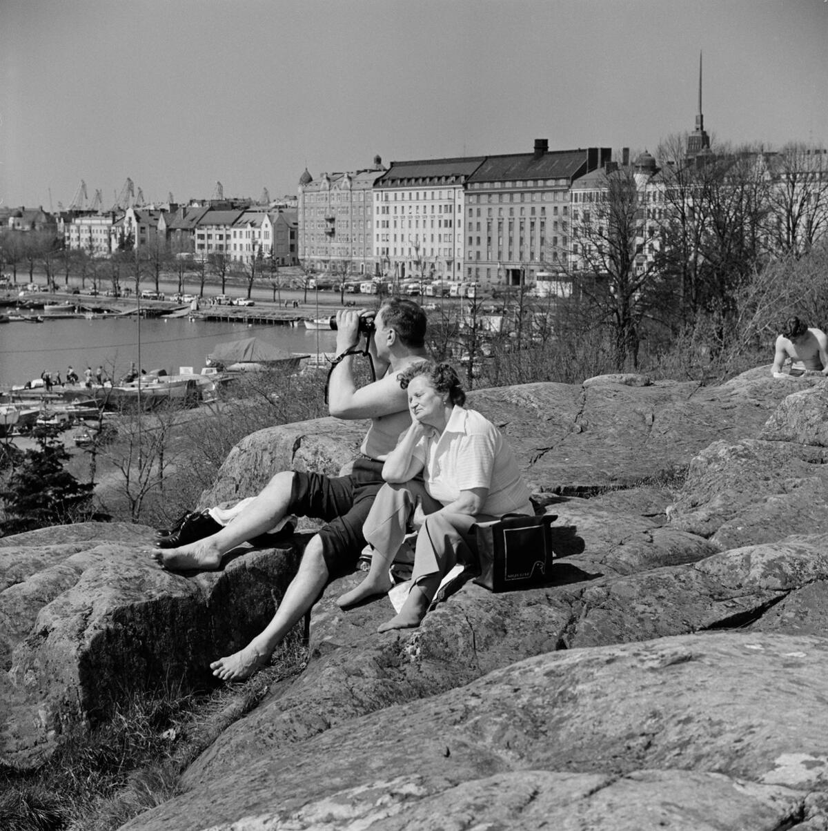 Pariskunta istuu Kaivopuiston kalliolla. Taustalla vasemmalla venerantaa, oikealla Merikatu.