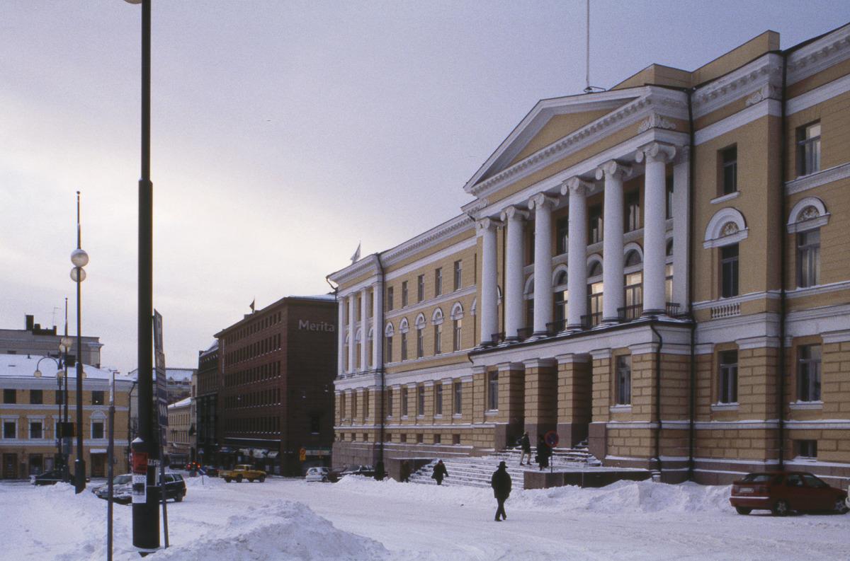 Etualalla näkyy keltaisen hallinnollisen rakennuksen julkisivu. Vuodenaika on talvi ja kadulla on lunta. Muutama henkilö käyskentelee rakennuksen portaiden tuntumassa.