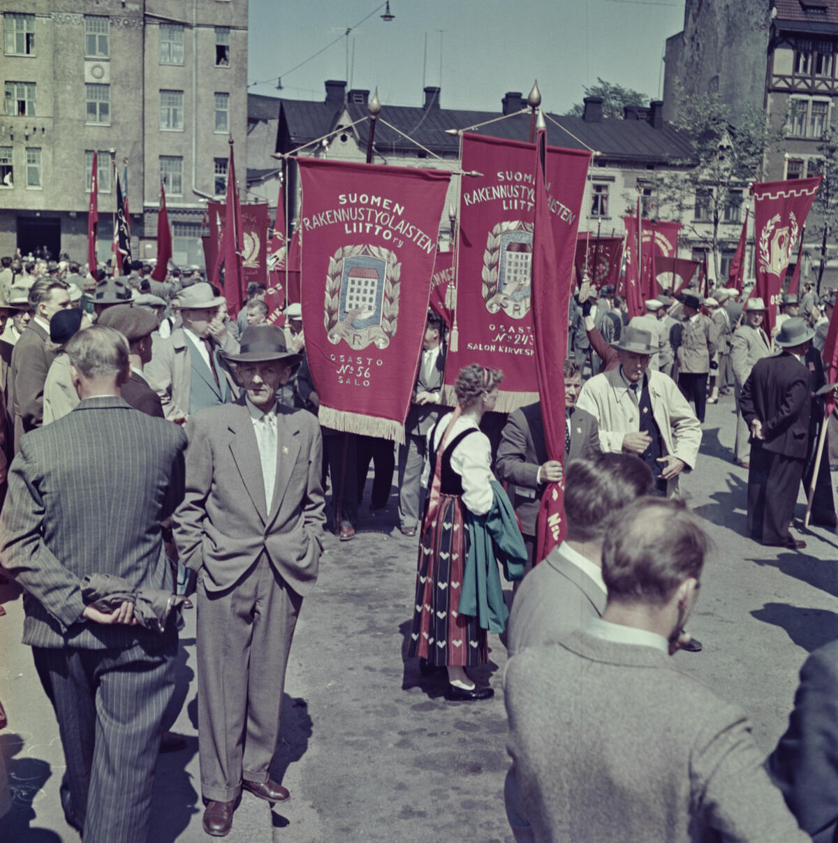Ihmisiä kokoontuneena Hakaniemen torille. Keskellä kuvaa on kansallispukuun pukeutunut nainen ja punaisia banderolleja, joissa teksti Suomen rakennustyöläisten liitto.