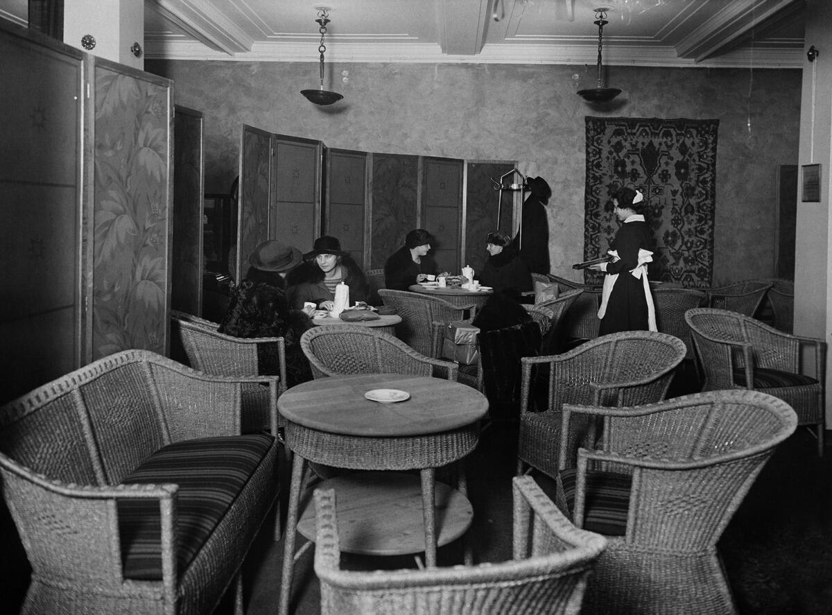 Korihuonekaluin kalustetussa kahvilassa istuu kahden pöydän ääressä kaksi naista, jolla on hattu päässä. Heitä kohti kävelee mustaan pukeutunut tarjoilija, jolla on valkoinen esiliina ja hilkka.