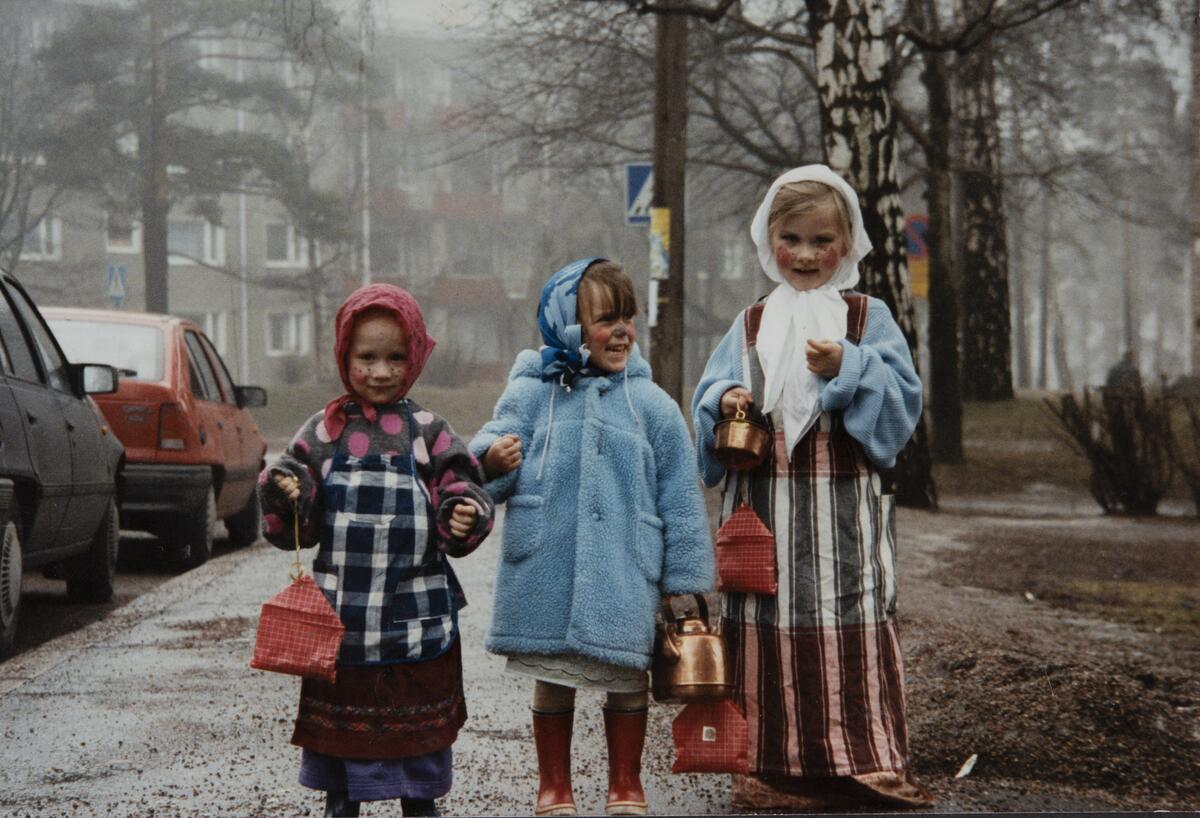 Kolme lasta autojen vieressä jalkakäytävällä pukeutuneina noidiksi, kupariset kahvipannut kädessä