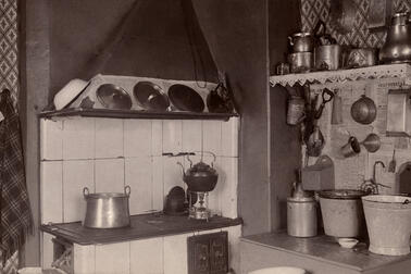 Vasemmalla on puuhella, oikealla on senkki, jonka päällä on sankoja. Seinällä on keittiövälineitä ja seinällä kattilahylly.