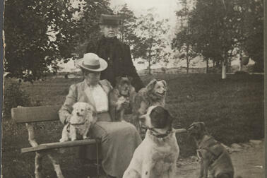 Kaksi naista puistossa. Toinen istuu penkillä, toinen seisoo hänen takanaan. Heidän kanssaan on viisi koiraa.