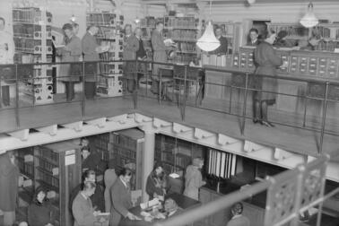 En vy mot två våningar i biblioteket på Rikhardsgatan. På den övre våningen studerar kunderna böcker och ett kartotek, på den nedre köar de till betjäningsdisken. 
