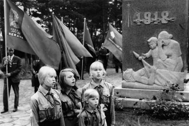 Neljä nuorta pioneeriasuista tyttöä laulaa Santahaminan veljeshaudalla punaisten muistomerkin edustalla. Taustalla lippuja piteleviä miehiä.