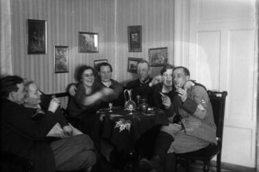 Seitsemän ihmistä istuu pyöreän pöydän ääressä. Joillakin on kädessä savuke ja snapsilasi. Pöydällä on snapsilaseja ja kaksi juomakaadinta. Yhdellä miehistä on suojeluskunnan univormu.