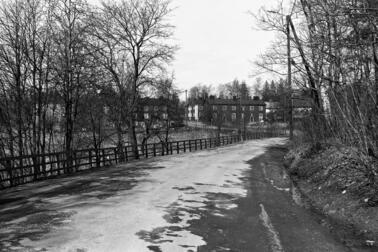 Maantie, jota reunustaa vasemmalla puukaide ja lehdettömät puut. Taustalla näkyy kaksikerroksisia puurakennuksia.