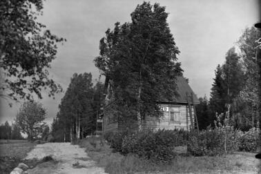 Vasemmalla on kapea hiekkatie, oikealla puolitoistakerroksinen puurakennus, jonka ympärillä on koivuja. Rakennuksen seinässä lukee ELANTO.