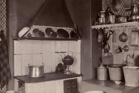 Vasemmalla on puuhella, oikealla on senkki, jonka päällä on sankoja. Seinällä on keittiövälineitä ja seinällä kattilahylly.