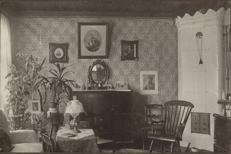 Huoneessa on kaakeliuuni, keinutuoli, kasveja sekä tauluja seinillä.