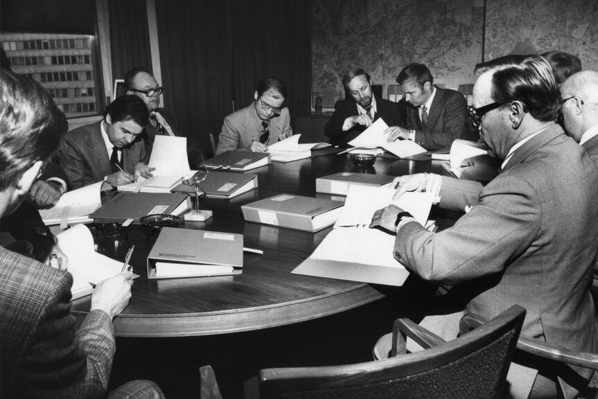 Helsingin metrotoimikunnan jäseniä kokouksessaan 1970-luvulla. Yhdeksän miestä istuu pöydän ympärillä tutkimassa asiakirjoja. Pöytä on täynnä paksuja mappeja.