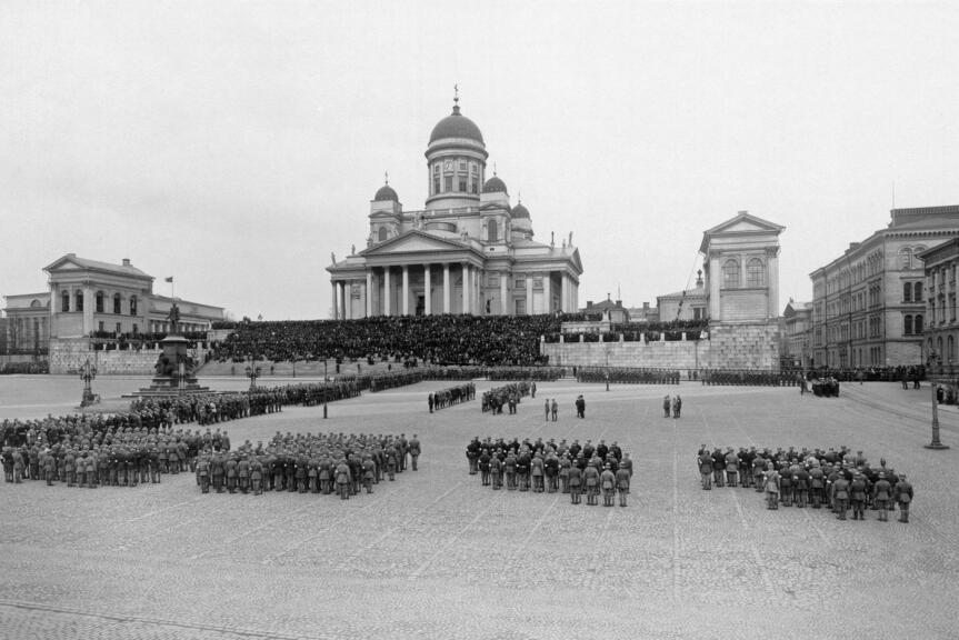 Jääkärit ovat järjestäytyneet paraatimuodostelmiin ympäri Senaatintoria. Ryhmittymien keskelle on kokoontunut oletettavasti jääkäriprikaatin päällystöä. Yleisö on kerääntynyt seuraamaan tapahtumia tuomiokirkon portaille.