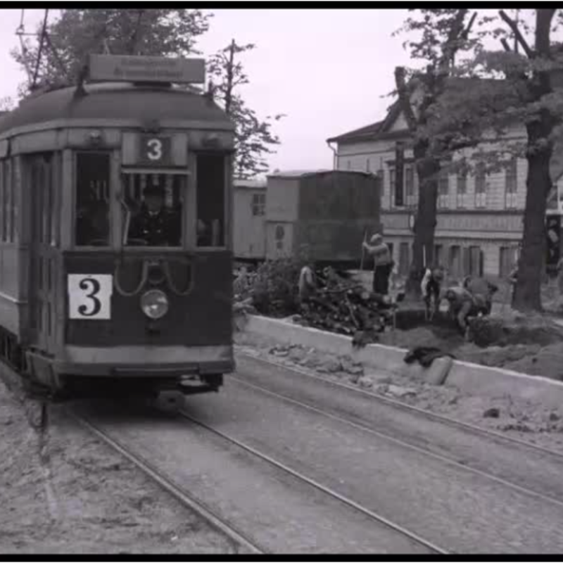 Vasemmalla on raitiovaunu kuvattuna edestä, oikealla työmiehiä rakentamassa Mannerheimintietä Vanhan ylioppilastalon kohdalla.