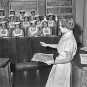 Ryhmä hoitajan työasuun pukeutuneita naisia istuu pienessä luentosalissa. Osa tekee muistiinpanoja. Heidän edessään sivuttain kuvaajaan seisoo nainen, jolla on vasemmassa kädessään paperi.