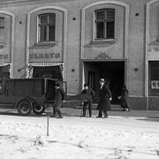 Luminen katu ja jalkakäytävä, jolle on pysähtynyt ambulanssi tai ruumiinkuljetusauto. Kolme miestä nostaa paareilla makaavaa, tummalla huovalla peitettyä ruumista auton takaosaan. Taustalla kävelee ohikulkija.