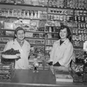 Kaksi hymyilevää naista seisoo kaupan tiskin takana. Toisella on tonttulakki päässään ja viinirypälevati kädessään. Naisten takana on hyllyjä täynnä ruokapakkauksia ja konvehtirasioita.