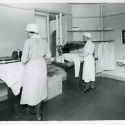 Kaksi valkoisiin työasuihin pukeutunutta naista seisoo selin sivuttain kameraan käyttämässä prässäyskonetta. Huoneessa on pari seinähyllyä ja pesuallas.
