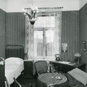 Vasemmalla on yhden hengen sänky, oikealla on kirjoituspöytä, jolla seisoo pöytälamppu ja puhelin. Keskellä huonetta on pieni pyöreä pöytä ja kaksi tuolia.