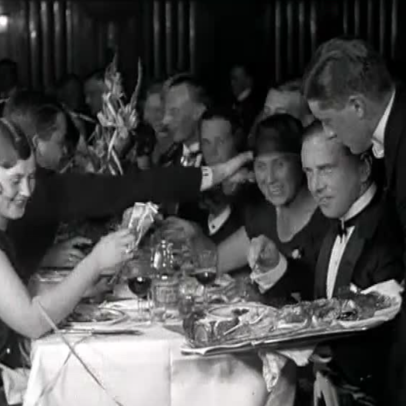 Juhlapukuisia nauravia ihmisiä pitkän illallispöydän ääressä. Miestarjoilija tarjoilee ruokaa vadilta.