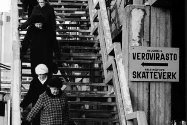 Ulkoportaissa kävelee viisi talvivaatteisiin pukeutunutta ihmistä. Portaat ovat aika jyrkät, ja jokainen pitää kiinni kaiteesta. Oikeassa reunassa näkyy Merihaan betonirakenteita.
