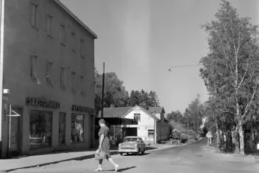 Mosabacka, Brödravägen 17–21. Till vänster finns ett rappat trevåningshus, vars bottenvåning utgörs av affärslokaler. På väggen hänger neonskyltar med texterna SÄÄSTöPANKKI och APTEEKKI APOTEK. En kvinna klädd i klänning går över vägen mot en byggnad. I bakgrunden finns en personbil och ett ljust trähus med sadeltak. Till höger finns björkar.