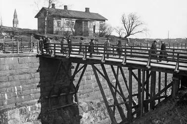 Människor promenerar på en träbro. Under bron löper hamnbanan. På andra sidan bron står en trävilla med en våning samt några kala björkar. I bildens vänstra kant syns Nationalmuseets torn.