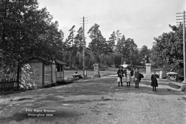 Stadsgräns, gammal tullport. Västra Chaussén 80 (numera Mannerheimvägen 108). Fyra barn står mitt på vägen, i bakgrunden syns en man som kör med häst. Till vänster om vägen finns en låg trästuga, och bakom den på båda sidorna om vägen finns stenpelare, som en tullbom tidigare vilat på.