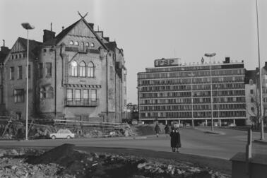 Rakennus purettiin pian kuvan ottamisen jälkeen, ja paikalle valmistui Ympyrätalo vuonna 1968. Kaksi ohikulkijaa on pysähtynyt katselemaan etualalla olevaa Kallion virastotalon rakennustyömaata. Oikealla näkyy Postisäästöpankin rakennus, Säästöpankinranta 2.