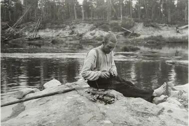 En man sitter på en klippa vid en å och öppnar musslor med en kniv. Bredvid honom är en hög öppnade musslor.