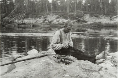 En man sitter på en klippa vid en å och öppnar musslor med en kniv. Bredvid honom är en hög öppnade musslor.