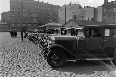 Vuokra-autoja siistissä rivissä mukulakivetyllä aukiolla. Taustalla vasemmalla kohoaa hotelli Seurahuoneen talo ja oikealla rakennuksia, jotka sijaitsivat ennen nykyisen Postitalon paikalla.
