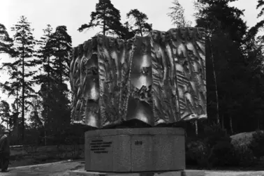 Kivipaadella lepäävä pronssinen muistomerkki, jossa abstraktin, litteän veistoksen poimuista työntyy esiin ihmispäitä.