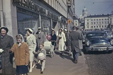 Människor med studentmössor promenerar på gatan. Södra kajen 18. I bildens högra kan en personbil av märket Buick och årsmodell 1954.